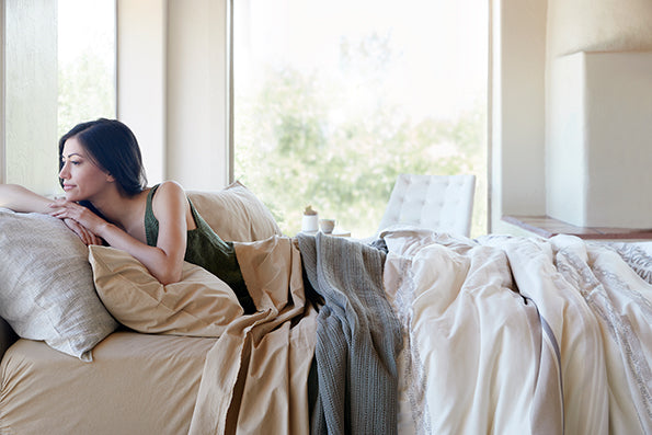 Image of a woman lying in bed, turned to look out the window behind her. The bed showcases Ochre Garment Washed Percale Sheets, an Agave Ridgeback Coverlet, and a Sonoran Duvet Cover.